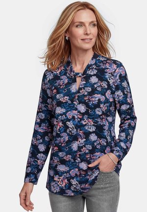 Femme aux cheveux blonds portant un chemisier bleu marine à manches longues à motif floral et un pantalon gris avec des détails en strass.