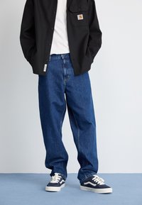 Blue denim wide-leg jeans with a button fly, paired with a white t-shirt and a black button-up shirt featuring a pocket. Navy sneakers.