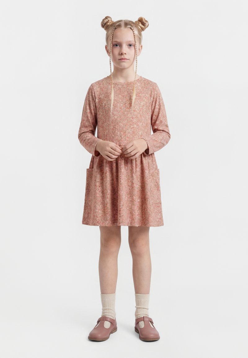 Jong meisje staat met het gezicht naar voren en haar handen samengevouwen, draagt een lange mouwen roze jurk met bloemenmotief, crèmekleurige sokken en roze Mary Jane schoenen.