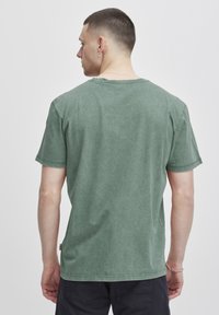Camiseta de algodón verde con un ajuste relajado, cuello redondo, mangas cortas y detalles de costura visibles. La vista trasera muestra una textura suave.