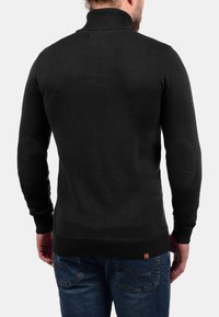 Schwarzer gestrickter Pullover mit hohem Kragen, gerippten Bündchen und Rückendetails. Mit Ellbogenflicken und eng anliegendem Schnitt. Wird mit blauen Jeans kombiniert.