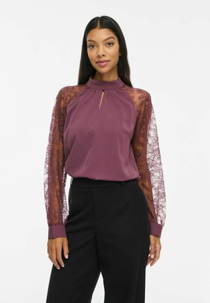 Une femme porte un chemisier couleur prune à manches en dentelle transparente et un pantalon noir, sur un fond blanc uni.
