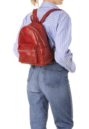 Sac à dos en cuir rouge de forme arrondie, avec une poche zippée à l'avant et des bretelles doubles, porté sur une chemise à rayures bleues.
