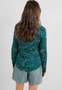 Blouse verte à motifs avec manches longues et taille cintrée ; présente de petits motifs d'oiseaux en blanc et beige ; portée avec un short gris clair.