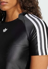 Svart atletisk topp med korta ärmar och glansig yta, med tre vita ränder på varje axel och en liten vit logga på bröstet.