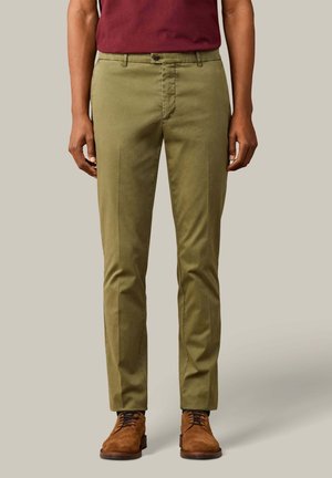 Hombre con pantalones ajustados verde oliva, zapatos de ante marrón con cordones y camisa burdeos, de pie frente a un fondo liso y claro.