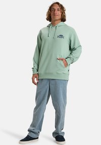 Sudadera verde menta con bolsillo tipo canguro y capucha con cordón, combinada con pantalones de corte relajado en azul claro y zapatos oscuros. Detalle del logo de Billabong.