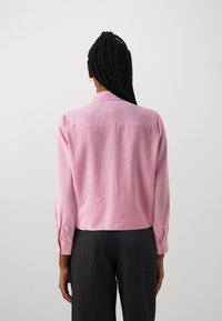 Camisa de pana rosa con un cuello estructurado, mangas largas y puños abotonados. Patrón de rayas verticales en la tela. Vista trasera.