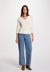 Cremfarbener Pullover mit V-Ausschnitt und Zickzackmuster, kombiniert mit hochgeschnittenen blauen Jeans und braunen Stiefeletten. Schlichter Hintergrund.
