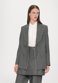 Grå skreddersydd blazer med dobbeltradet design, kontrastsømmer og runde reverser. Brukt over en hvit skjorte med krage.
