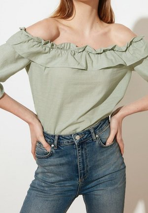 Mujer con blusa verde claro de hombros descubiertos y volantes, metida en jeans azules de tiro alto, con las manos apoyadas en las caderas.