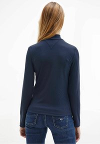 Marinblå långärmad topp med en texturerad design, med en V-formad halsringning och en figurnära siluett. Matchas med blå denimjeans.