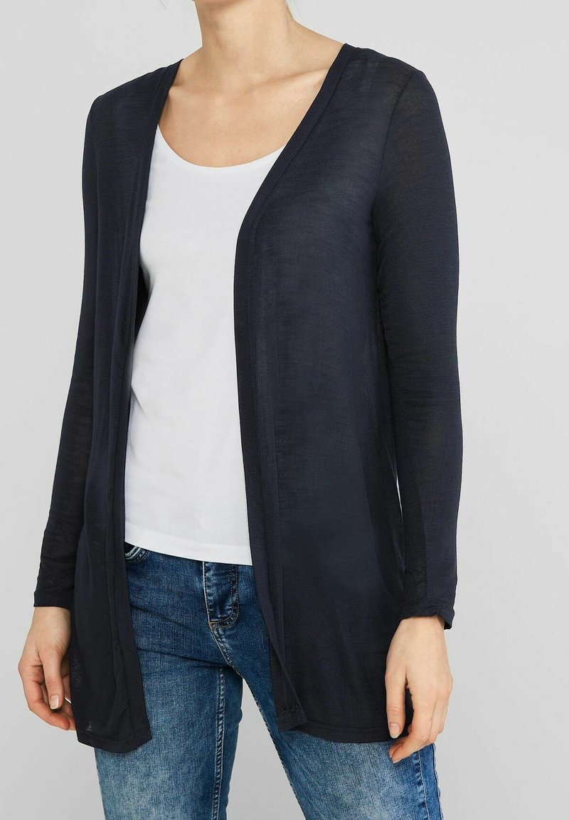 Žena v průhledném černém kardiganu s otevřeným předním dílem, pod kterým má bílé triko s výstřihem ve tvaru lodě, a modrými džíny, stojí na jednobarevném světlém pozadí.