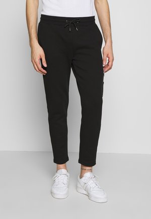 Homme portant un pantalon de jogging noir slim avec taille à cordon et des baskets blanches, debout sur un fond gris uni.
