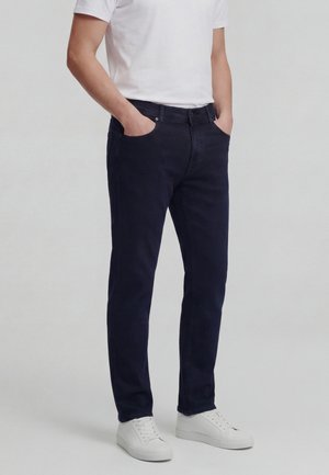 Mand iført mørkeblå jeans, hvid t-shirt og hvide sneakers, stående med hænderne i lommerne mod en ensfarvet baggrund.