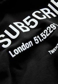 Fekete szövet, amelyen merész, fehér szöveg olvasható: "SUB5CRIBE London 51.5229°", puha textúrával és látható minták vagy hangsúlyok nélkül.
