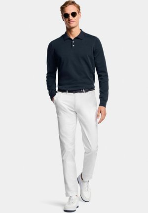 Homme portant un polo noir à manches longues, un pantalon blanc, des baskets blanches et des lunettes de soleil, marchant vers l'avant avec une main dans la poche.