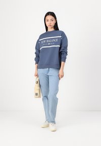 Navy sweatshirt with white "NEW BALANCE" logo, relaxed fit, and ribbed cuffs; paired with light blue jeans and white sneakers. Carrying a beige bag.