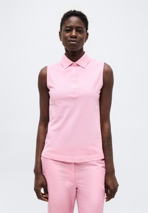 Femme aux cheveux courts et bouclés portant un polo rose sans manches et un pantalon rose assorti, debout sur un fond blanc.