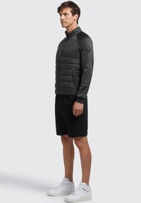 Men's black puffer jacket with a smooth, fitted fabric and zipped pockets, paired with black shorts and white high-top sneakers.
