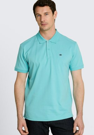 Mężczyzna w jasnoniebieskiej koszulce polo z krótkim rękawem, kołnierzykiem i dwoma guzikami, stojący na jasnym, jednolitym tle.