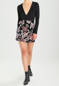 Bodysuit noir à manches longues avec un décolleté en V profond, associé à une jupe noire courte ornée de broderies florales colorées. Bottines noires à la cheville.