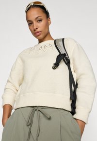 Sudadera de felpa color crema con costuras decorativas en forma de diamante. Combinada con pantalones verdes de cintura ajustable con cordón y un bolso de tirante negro.