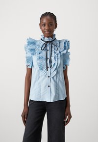 Gaiši zila blūze ar viļņotām piedurknēm, augstu apkakli un pogu aizdari priekšā; satur melnu kakla lentu un krokotu tekstūru.