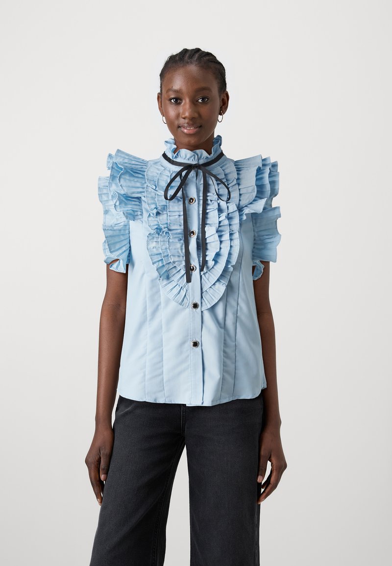 Gaiši zila blūze ar viļņotām piedurknēm, augstu apkakli un pogu aizdari priekšā; satur melnu kakla lentu un krokotu tekstūru.