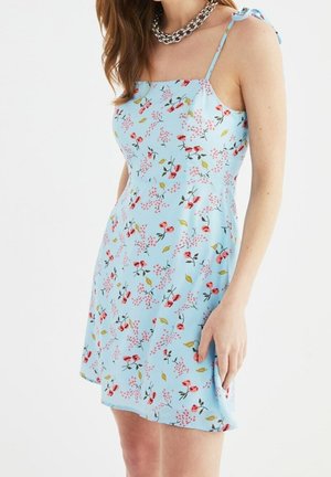 Frau trägt ein hellblaues, ärmelloses Kleid mit Blumenmuster, dünnen Trägern, die an den Schultern gebunden sind, und eine silberne Ketten-Halskette.