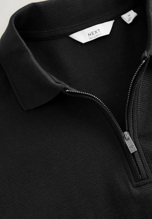 Sort struktureret polo-shirt med en sølvfarvet halvlukning og hvide mærker med teksten "NEXT Regular Fit" og størrelse "M."