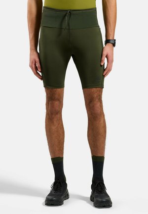 Mann trägt olivgrüne Sportshorts, schwarze Socken und schwarze Laufschuhe, steht mit entspannten Armen seitlich vor weißem Hintergrund.