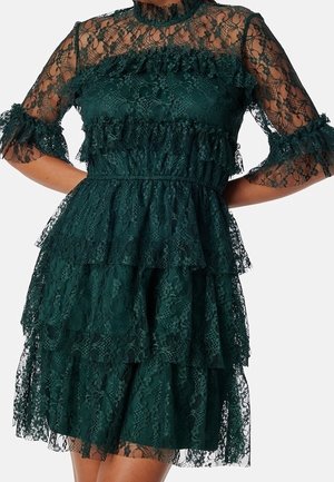 Robe en dentelle vert foncé avec volants superposés, manches en dentelle transparente et col montant, cintrée à la taille, portée par une personne les mains sur les hanches.
