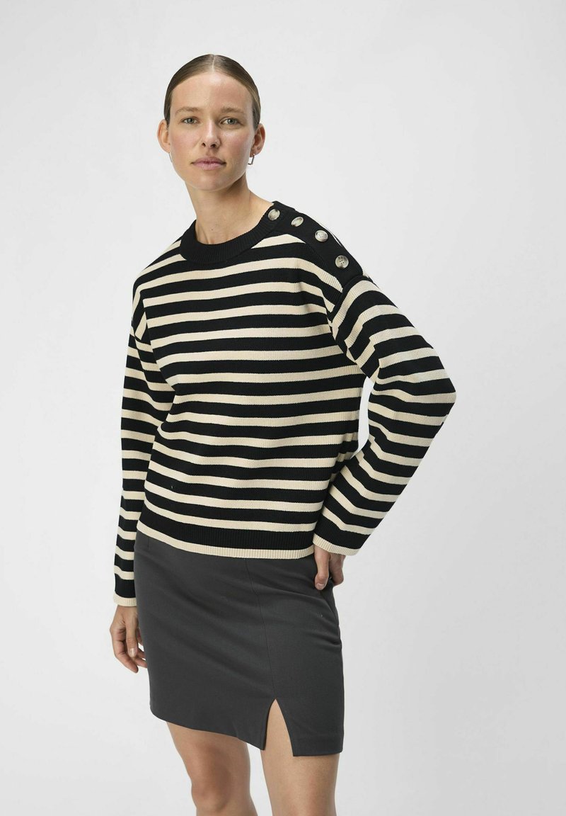 Gestreifter Strickpullover in Schwarz und Creme mit Rundhalsausschnitt und Knopfdetails auf der Schulter, kombiniert mit einem schwarzen Minirock mit seitlichem Schlitz.
