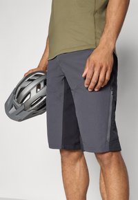 Shorts gris avec une poche zippée, fabriqués à partir d'un matériau léger. Chemise à manches courtes olive, tenue avec un casque de vélo gris.