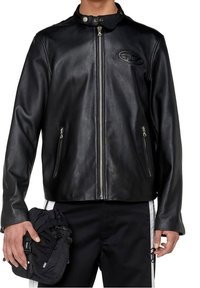 Uomo che indossa una giacca nera in pelle con zip e tasche con zip, tiene in una mano una borsa nera con cordino e indossa pantaloni neri con strisce bianche.