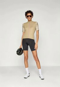 Jersey de ciclismo beige con mangas cortas y una cremallera rosa, combinado con pantalones cortos negros. El modelo sostiene un casco negro y lleva calcetines y zapatos blancos.