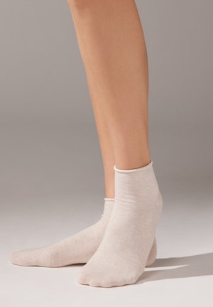 Par de piernas desnudas con calcetines tobilleros de color beige claro, de pie sobre una superficie lisa de color neutro con un fondo sencillo.