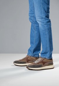 Braune und graue Turnschuhe mit strukturiertem Mesh und Leder, ausgestattet mit weißen Schnürsenkeln und einer dicken, cremefarbenen Sohle, getragen zu blauen Jeans.