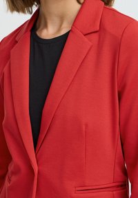Blazer rosso in tessuto a maglia testurizzata, con colletto a punte e tasche laterali, sovrapposto a un top nero. Design pulito e sartoriale.