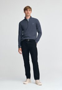 Mörkgrå zip-up-tröja med lång ärm, bärs med svarta byxor och ljusfärgade sneakers. Har en liten logotyp på bröstet.