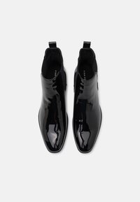Botas de tobillo de charol negro con un diseño elegante, punteras puntiagudas y paneles laterales elásticos para facilitar su uso. Incluyen una asa en el talón.