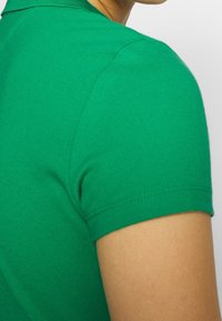 Vihreä t-paita lyhyillä hihoilla, jossa on ribatut kaulukset ja sileä kangas. Näkyvää ompelua hiha-aukossa.