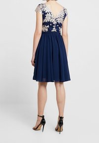 Marinblå klänning med en figursydd spetsöverdel som har crèmefärgad broderi och en veckad chiffongkjol, tillsammans med svarta högklackade sandaler.