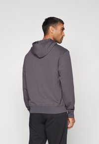 Felpa grigia con cappuccio regolabile, polsini e orlo a costine. Tessuto liscio e vestibilità casual, abbinata a pantaloni della tuta scuri.