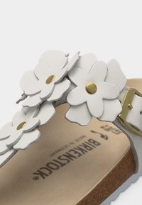 Λευκό λουράκι σανδάλου με διακοσμητικά λευκά λουλούδια με χρυσά κέντρα, μπεζ σόλα φελλού και χρυσό κούμπωμα.
