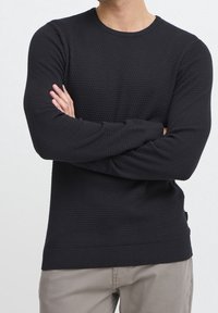 Maglione nero in maglia con texture, maniche lunghe e polsini a costine. Presenta un collo rotondo e un dettaglio con etichetta laterale. Indossato sopra dei pantaloni chiari.