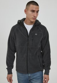 Grau fleece Jacke mit Kapuze, Reißverschluss vorne und Brusttasche. Verfügt über eine weiche Textur und gerippte Bündchen, getragen über einem weißen T-Shirt.