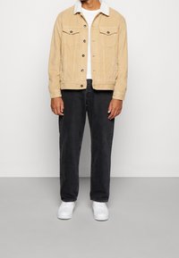 Beige manchesterjacka med fårskinnskrage, framfickor och svarta jeans. Modellen bär vita sneakers. Enkel, strukturerad design.