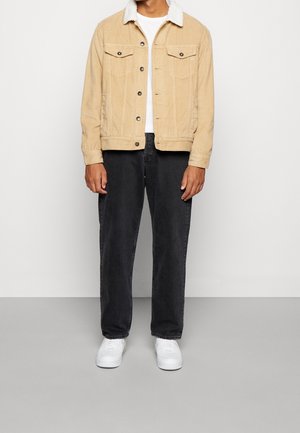 Beżowa kurtka sztruksowa z kołnierzem z kożucha, przednimi kieszeniami i czarne dżinsy. Model nosi białe trampki. Prosty, strukturalny fason.